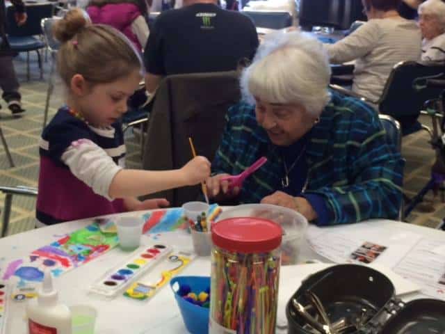 A woman helps a young child as she does arts and crafts; together they smile as they paint and cut out paper.