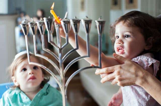 An image of a young girl, with help from an adult, lighting and placing the candles on a menorah. A boy even younger than her, watches in both amazement and slight fear.