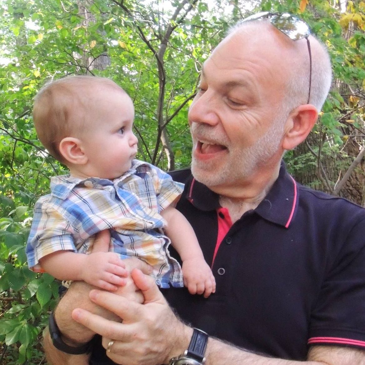 An older man with a collared shirt and glasses on his head holds a baby, smiling and talking to him as the baby stares back at the man in awe.