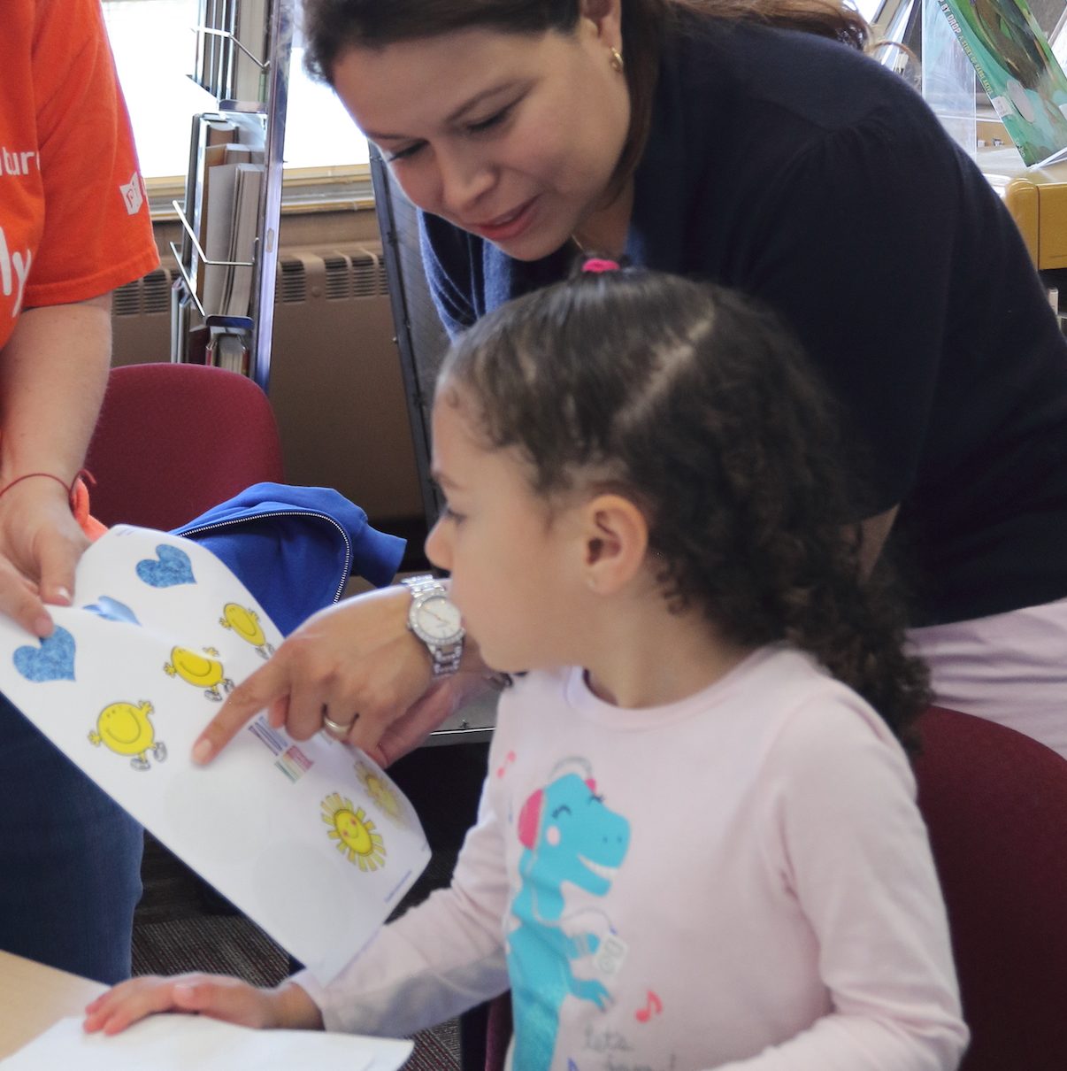 A woman teaches a young girl, as they both look at a piece of paper with yellow and blue cartoon figures on it. The girl is engaged in what she is learning as she turns to see what the woman is pointing at on the page.