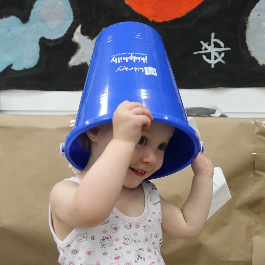 A young child peeks through a blue bucket that she has on her head, with the words “jkidphilly” written on it. She is playful, happy, and smiling.