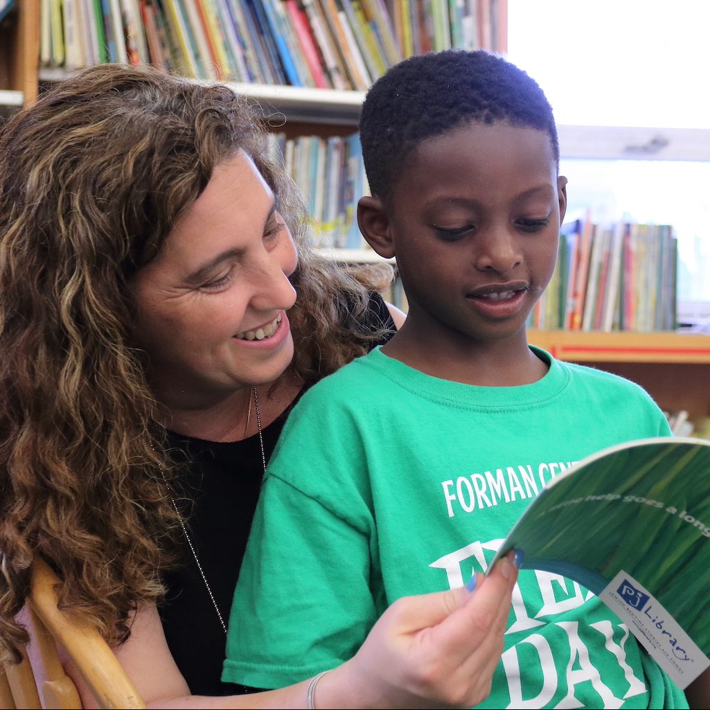 A woman smiling as she sits behind a young boy and reads with him. They are both happy and enjoying each other’s company as well as the book.