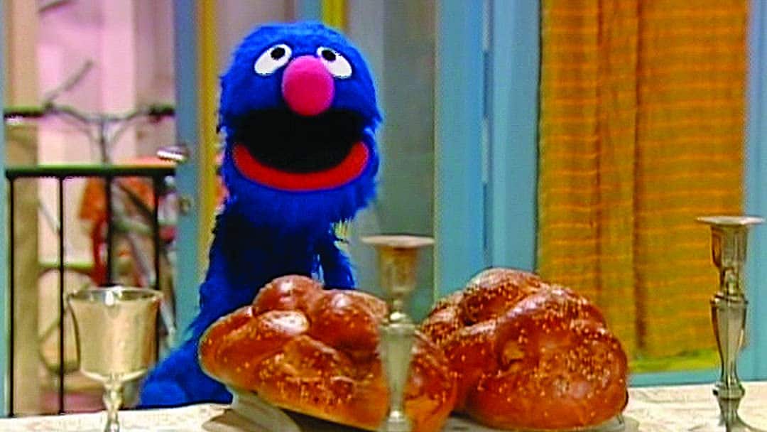 An image of a character on sesame street celebrating shabbat with challah and candles.