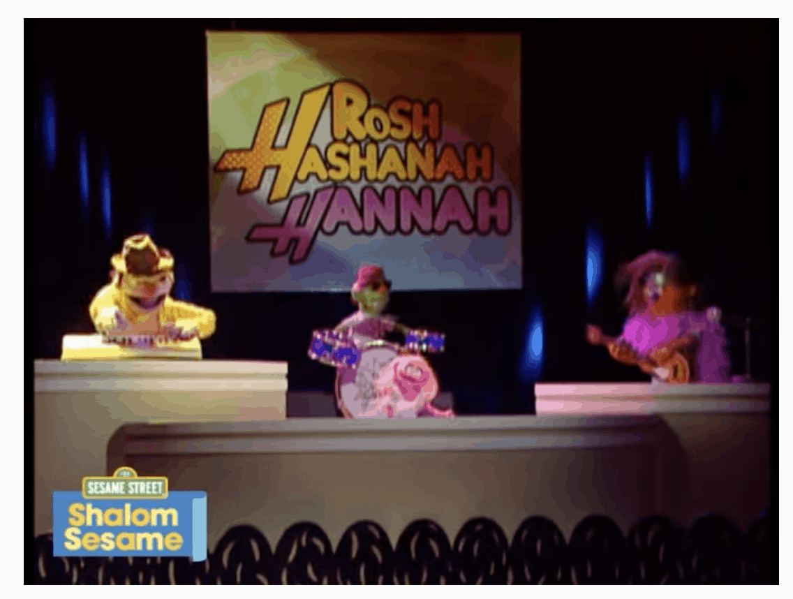 A screenshot of a Shalom Sesame video. Three muppets are on a stage playing music, with a big sign in the background that says "Rosh Hashanah Hannah," in a font reminiscent of Hannah Montana.