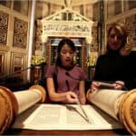 A photograph of an Asian teenager (named Cece Nealon-Shapiro) with long black hair and a purple dress reading from the Torah in front of an elaborate ark. 