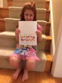 A photograph of a child sitting on carpeted stairs holding a colored sign that says 'welcome' in Hebrew