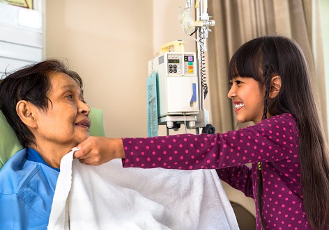 A photograph of a child adjusting a relative's blanket in a hospital. They are both smiling at each other.