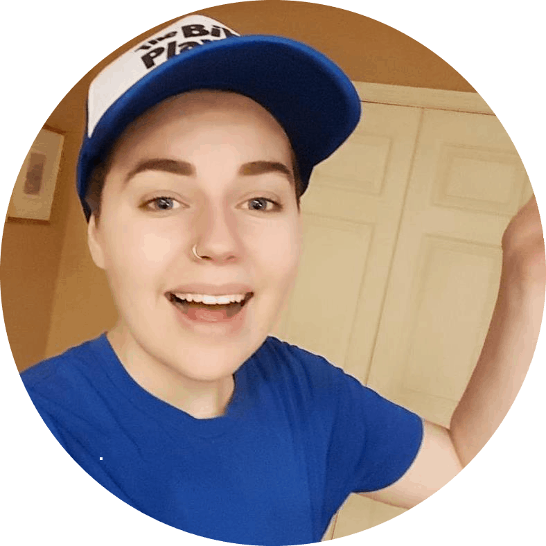 A photograph of a white non-binary person wearing a blue t-shirt and a white baseball cap. They are looking at the camera and seem to be in the middle of speaking while smiling.