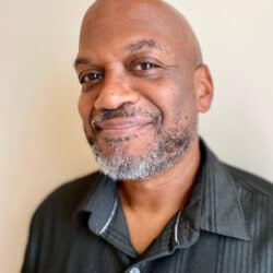 The headshot of a bald Black man with a greying beard and a charcoal button-up shirt. He is smiling warmly.