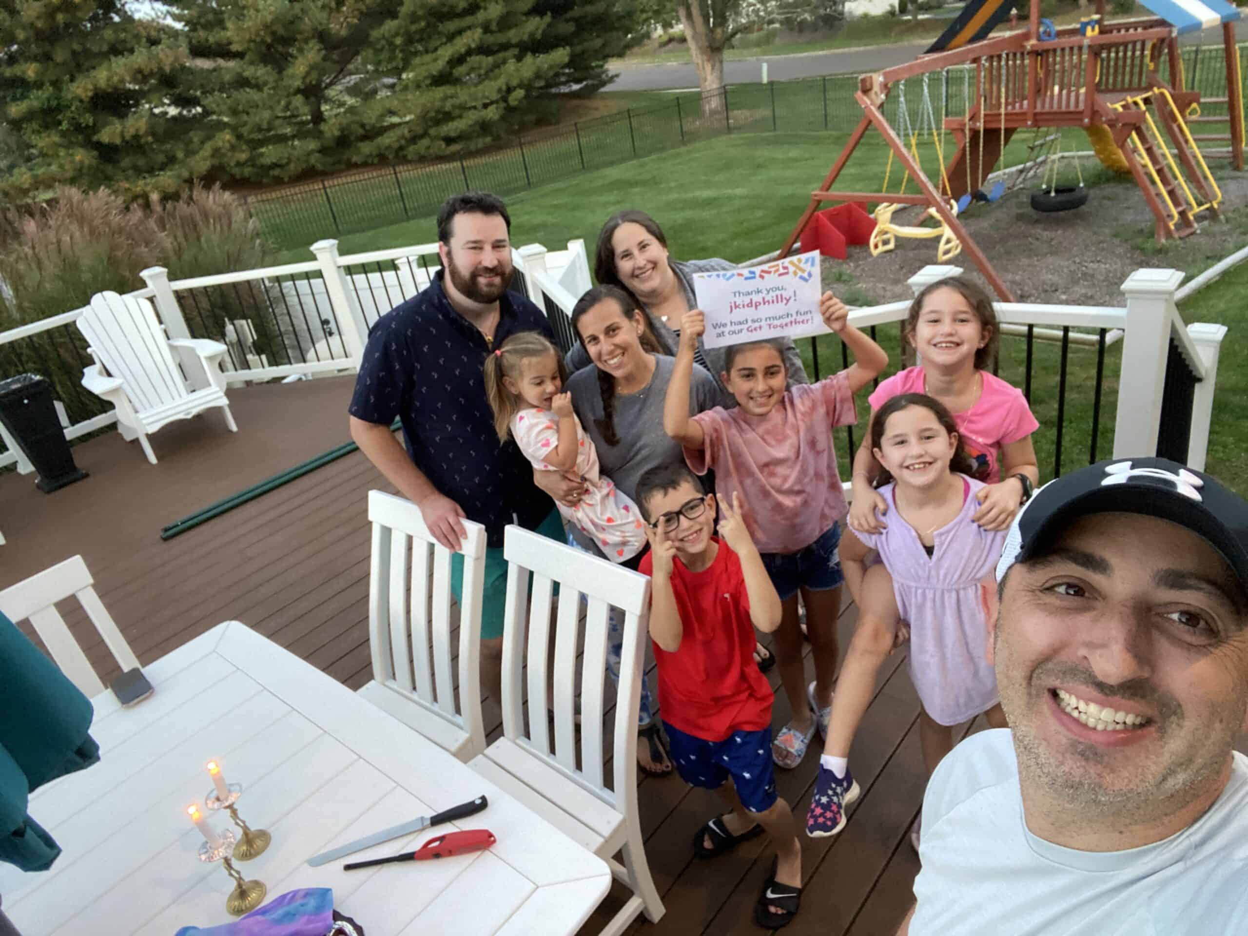 A photo of a large group of adults and children standing on a back porch, holding a sign that says thanks jkidphilly we had so much fun at our get together