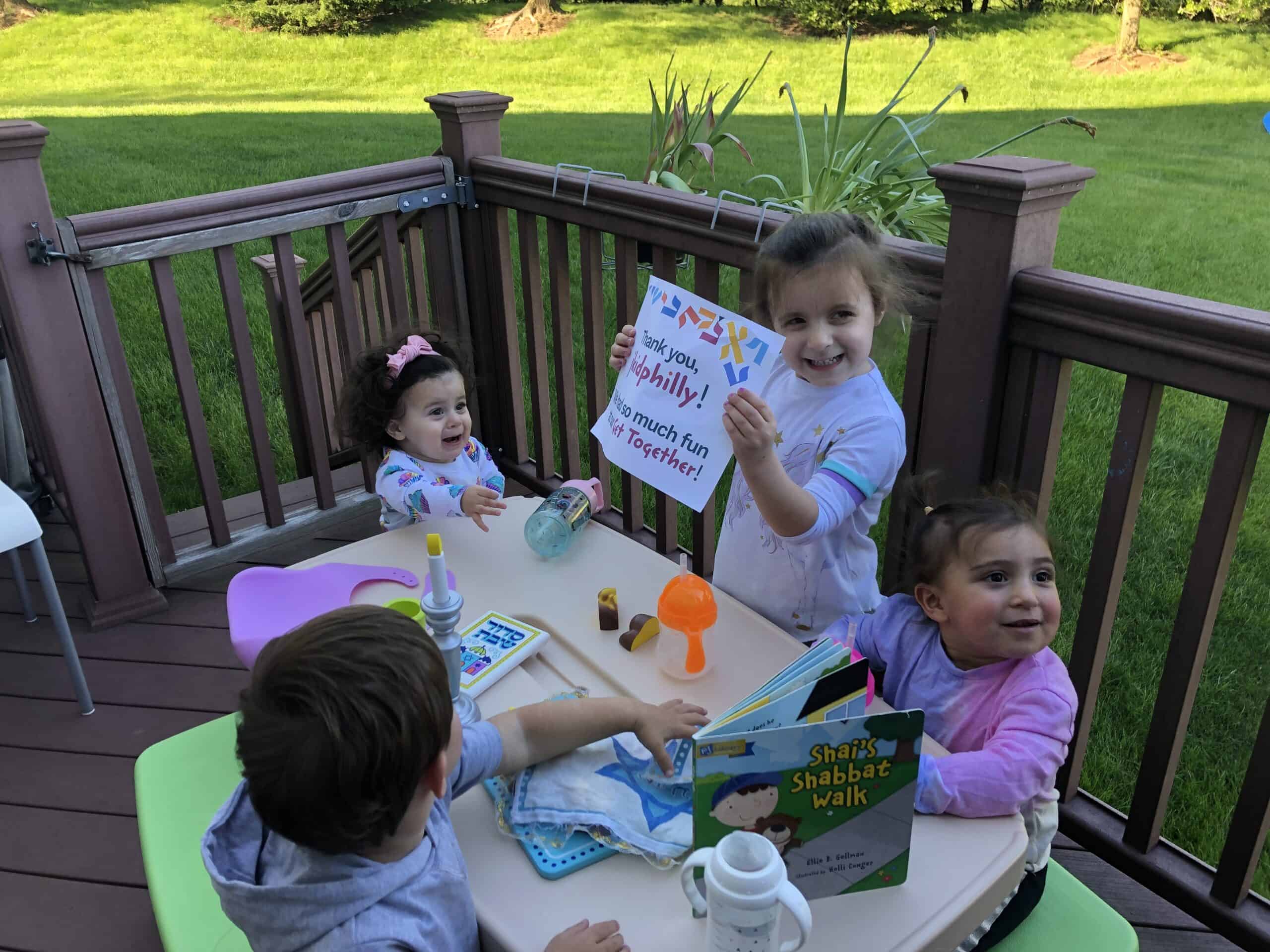 A photo of four toddlers sitting around a table covered in toy versions of Shabbat supplies, next to the book Shai's Shabbat Walk. One of the toddlers is holding a sign that says, Thanks jkidphilly, we had so much fun at our Get Together.