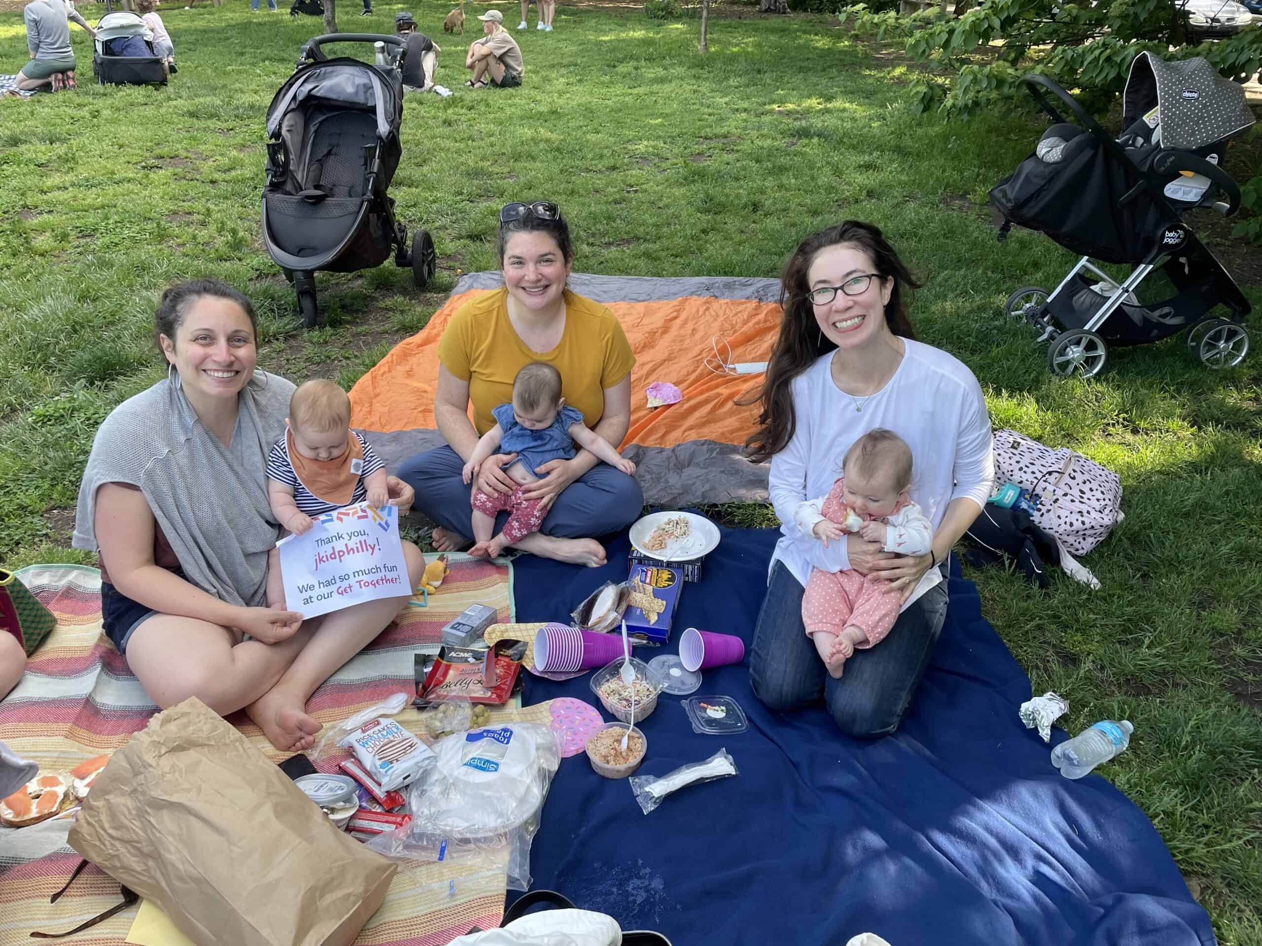 A photo of three adults holding three babies while sitting on a picnic blanket