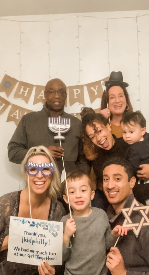 A photo of several adults and small children in front of a sign that says Happy Hanukkah. Several of the adults are holding cardboard cutouts of Hanukkah imagery. One is holding a sign saying, Thanks jkidphilly, we had so much fun at our Get Together.