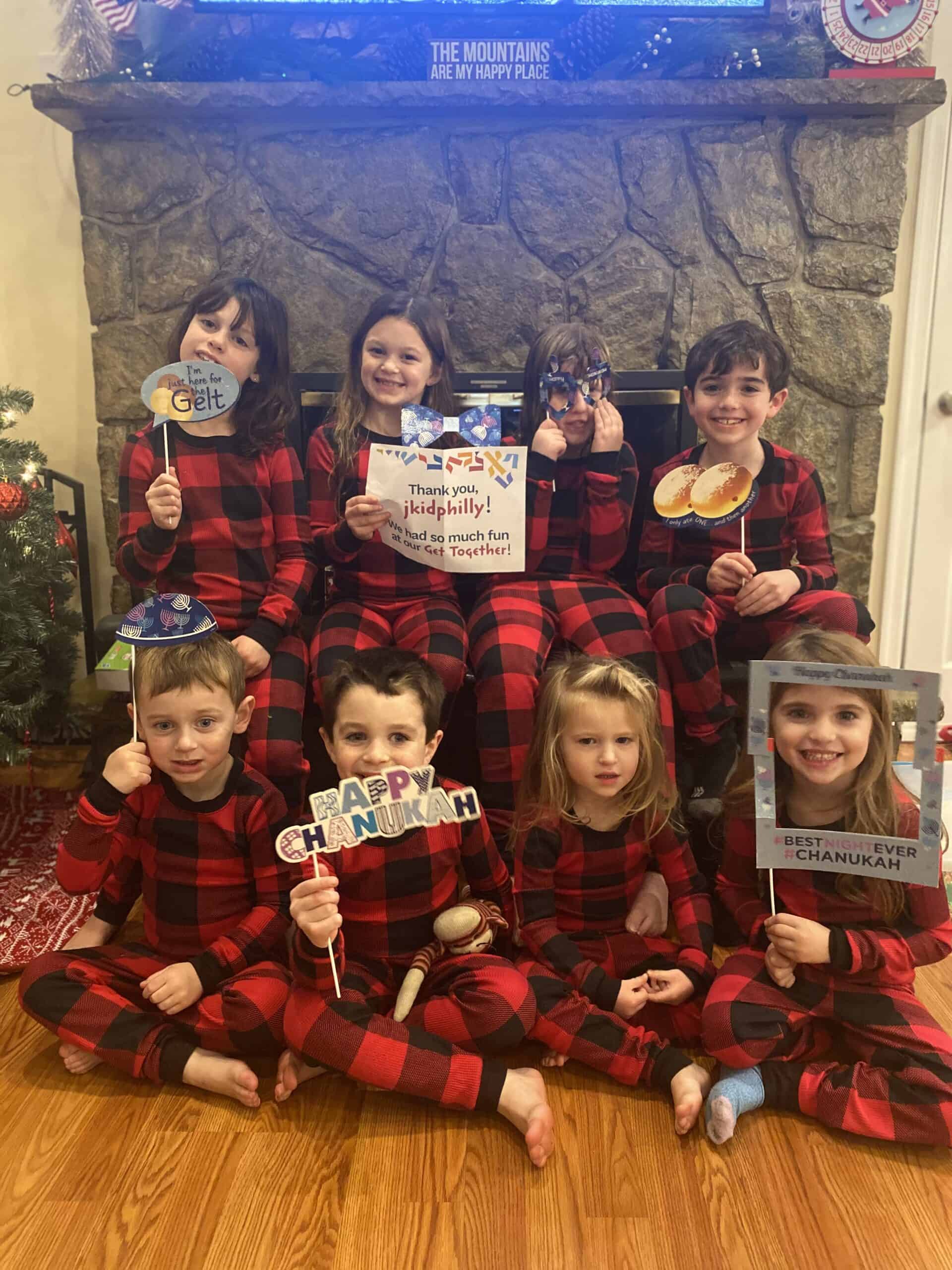 A photo of eight children, all in matching red plaid pajamas, holding selfie props relating to hanukkah
