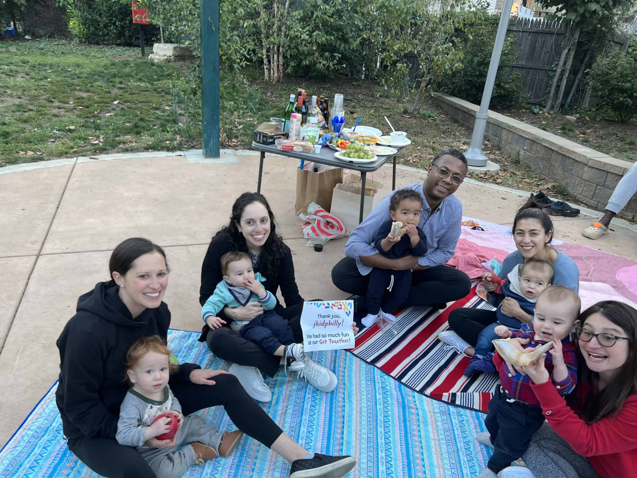 A phot of several adults, each with a baby in their lap, sitting on picnic blankets holding a sign that says thank you jkidphilly we had so much fun at our get together