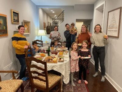 A photo of a large group of adults, adolescents, and children smiling and standing around a table covered with food.