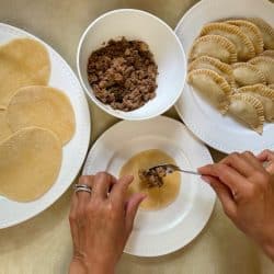 A photo of someone filling an empanada wrapper with brisket filling, next to a plate of filled empanadas