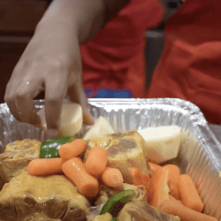 A large foil tray of lamb and vegetables, with someone with tongs mixing it.