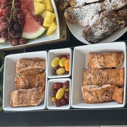 A photo of several plates of grilled salmon on a table with other plates with fruit, bread, and olives.